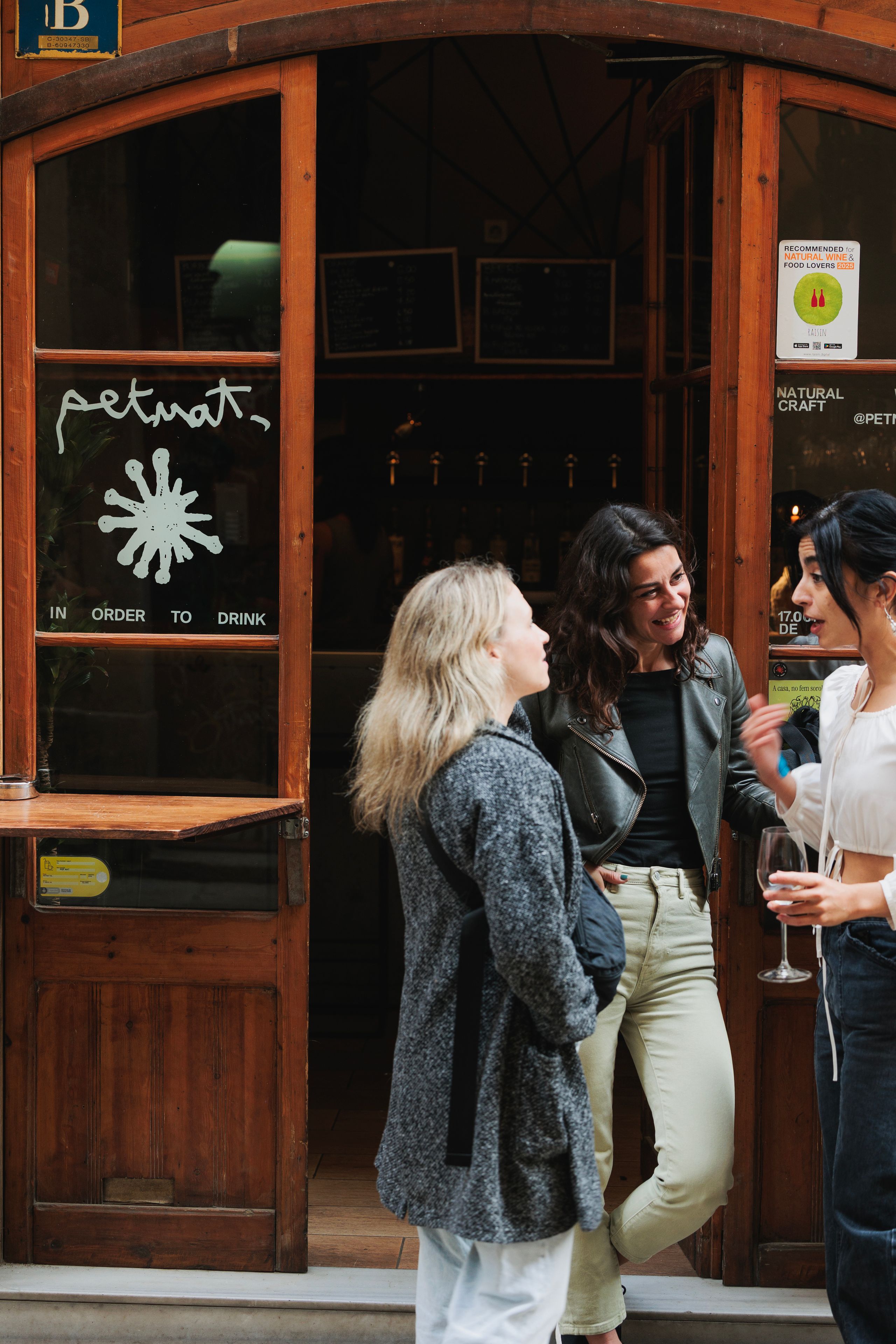 Three adults standing and chatting outside a café or bar with wooden doors; one person holds a wine glass while they laugh and talk together. Barcelona Neighbourhood