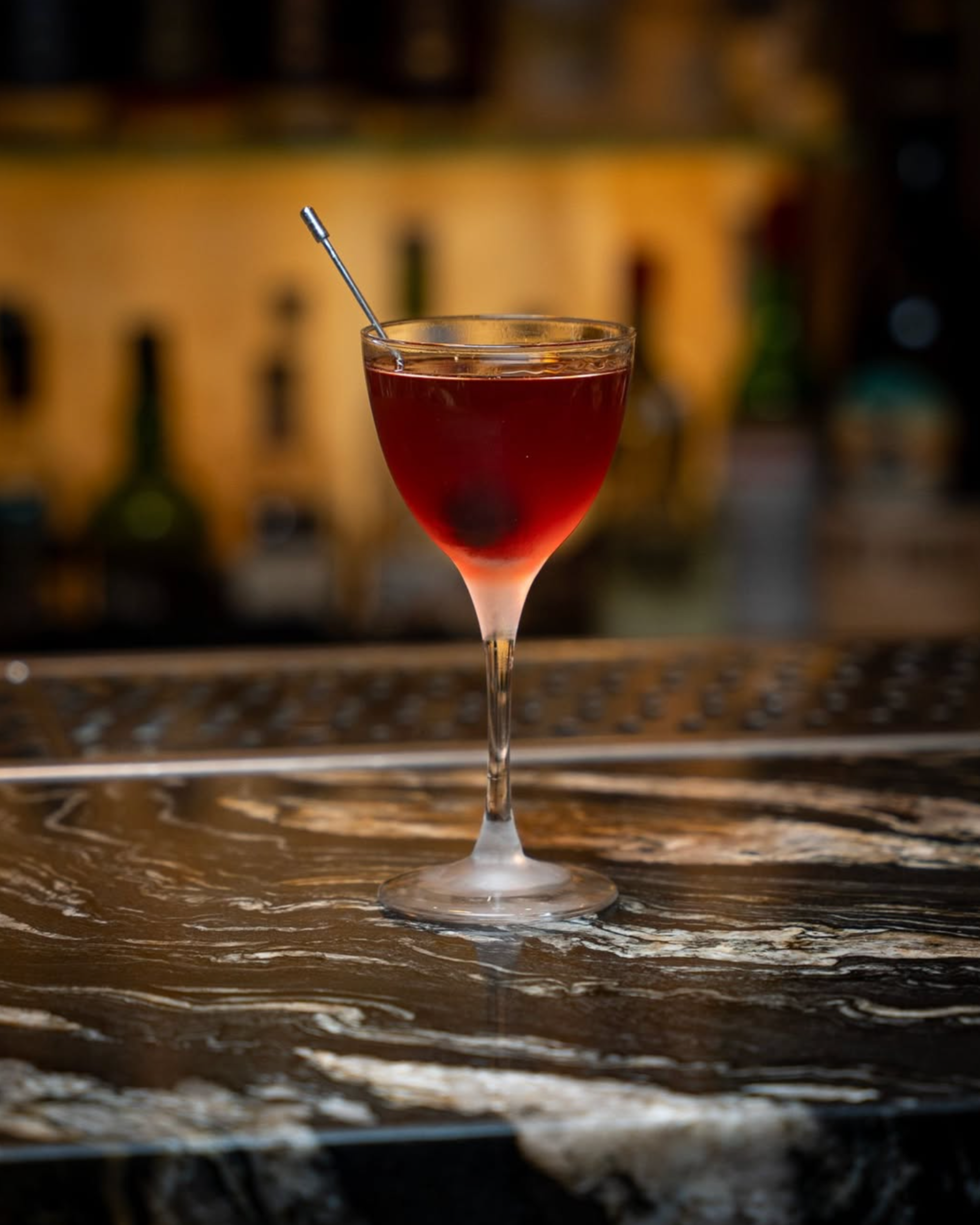 Elegant red cocktail in a stemmed glass with a metal stirrer on a marble bar surface.