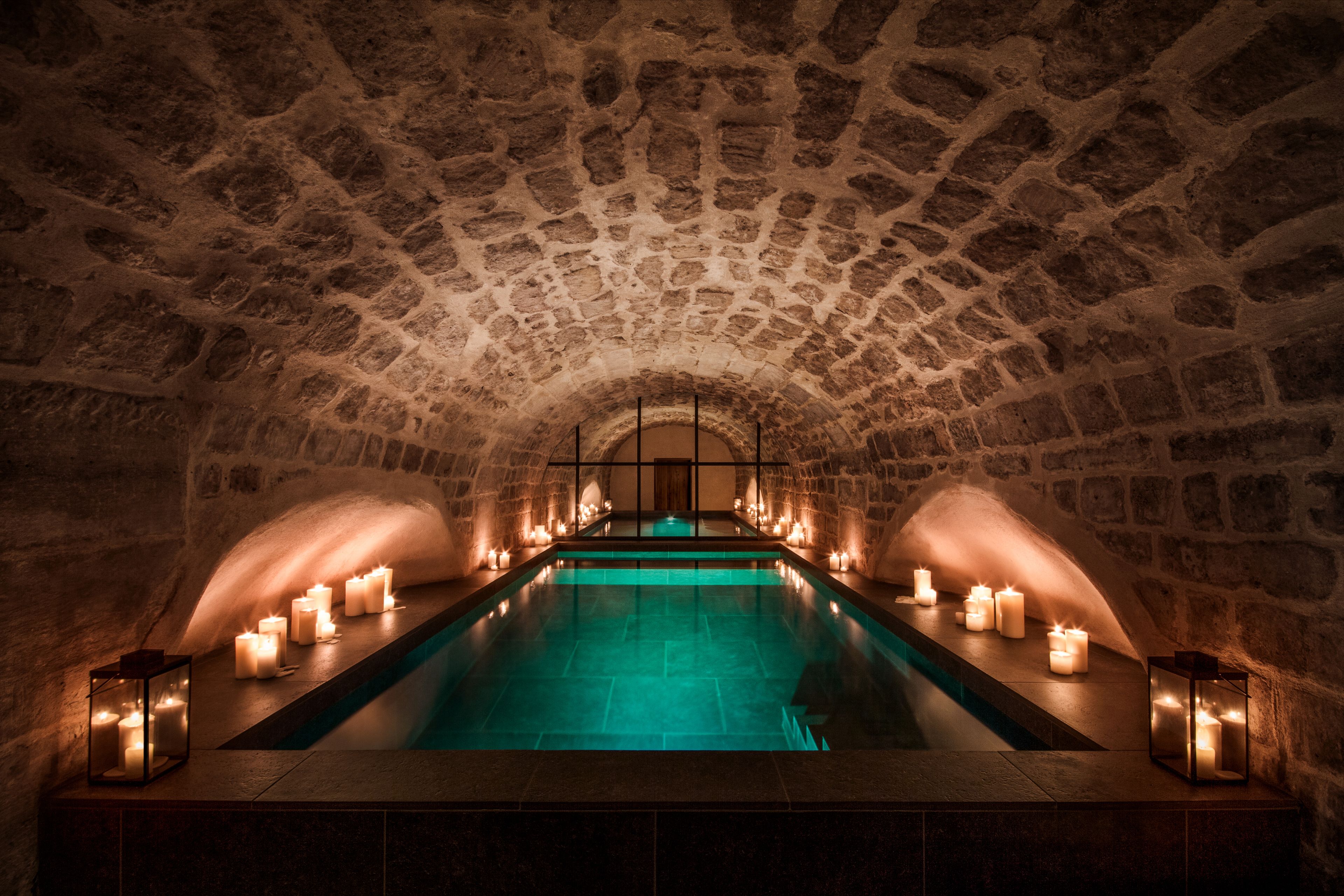 A dimly lit, elegant indoor pool set beneath a vaulted stone ceiling, with glowing candles placed along the pool’s edge and in lanterns, creating a warm, serene spa-like atmosphere.