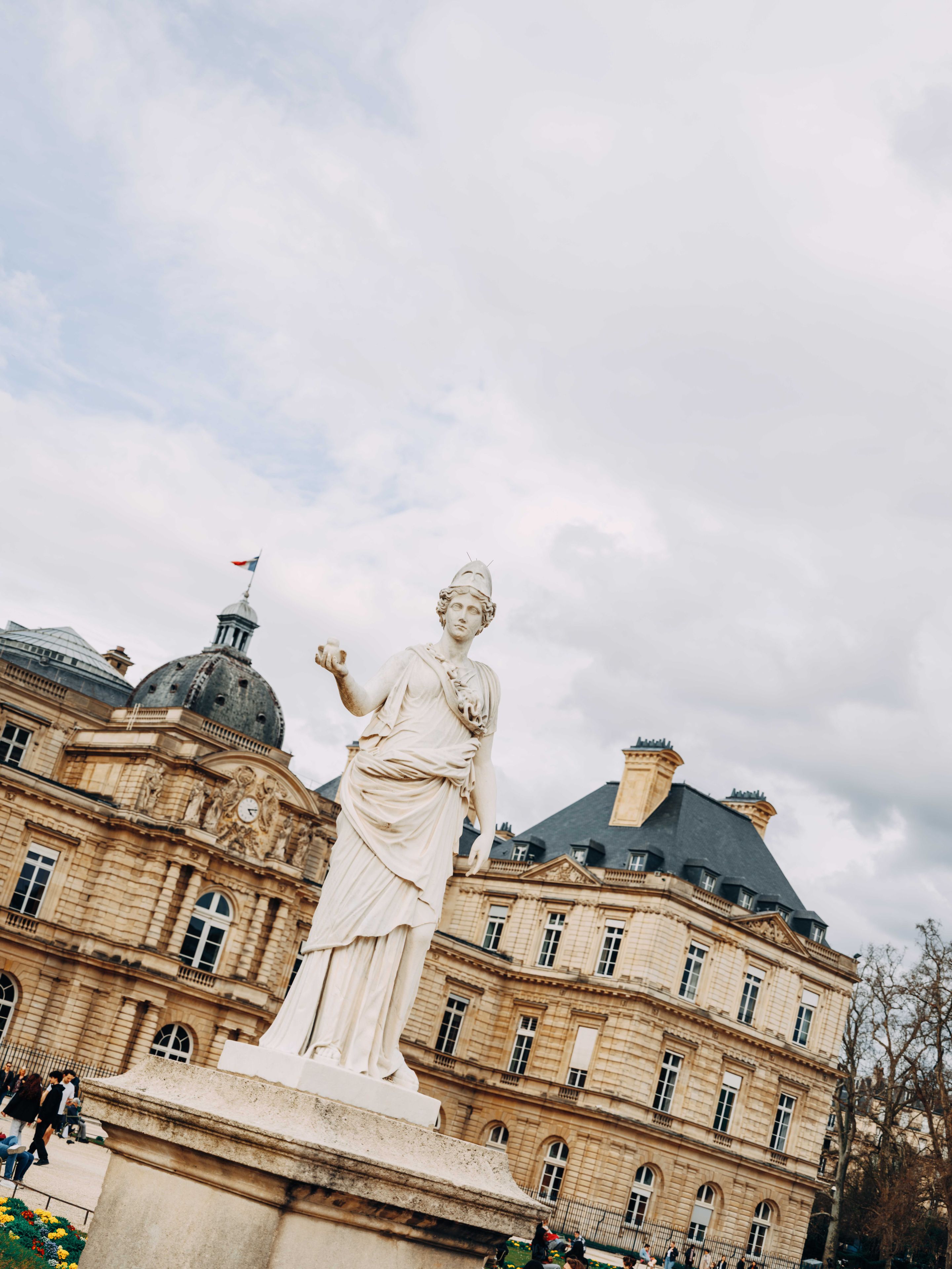 A carved marble statue of a classical figure in flowing robes stands atop a pedestal in the Jardin du Luxembourg in Paris, with the elegant stone architecture of the Luxembourg Palace and its dome in the background under a cloudy sky.