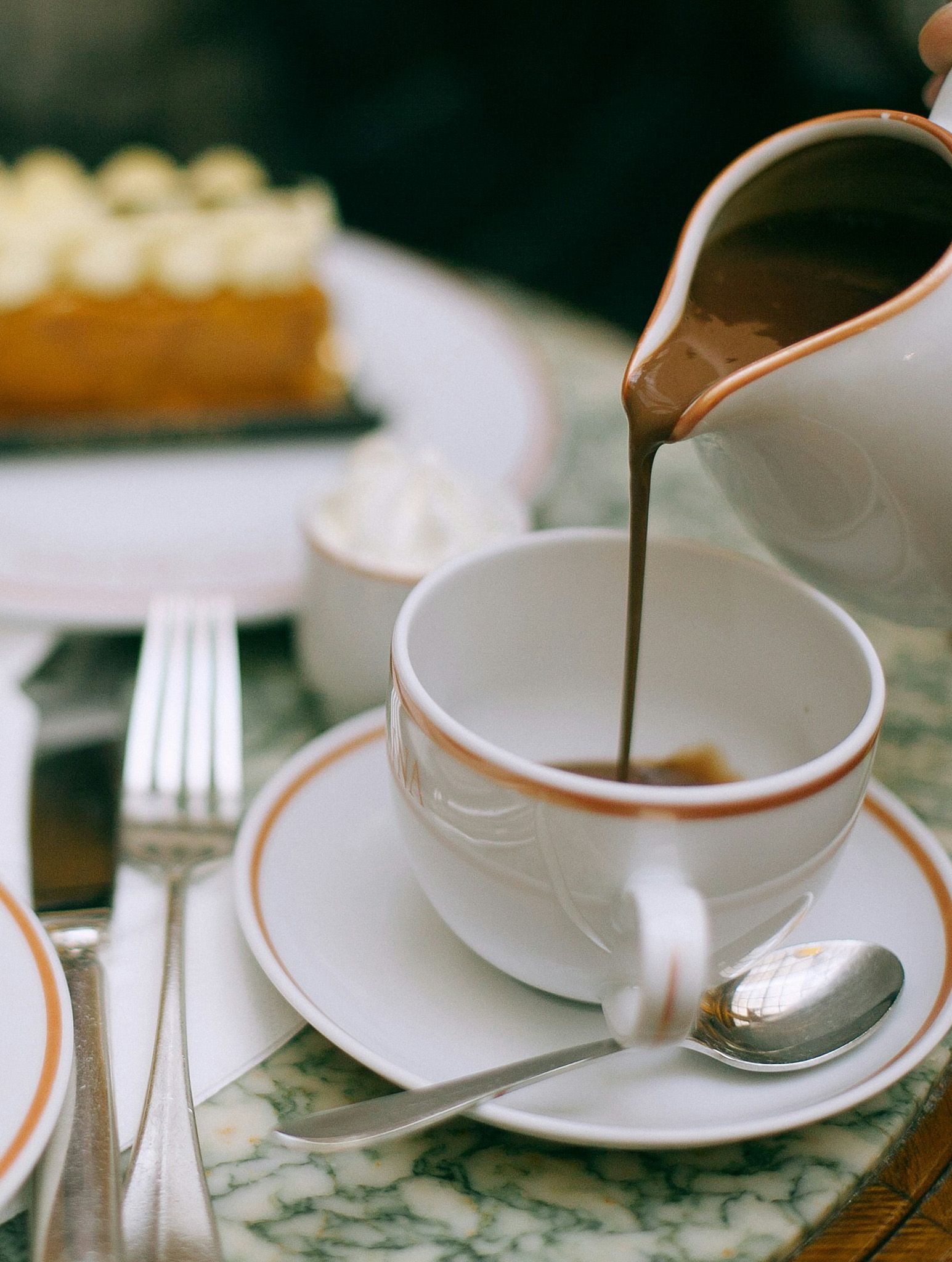 A close-up of a white porcelain cup filled with rich hot chocolate as a server pours more chocolate from a small pitcher, creating a smooth, glossy stream. The scene captures a warm, indulgent moment in an elegant Paris café setting.