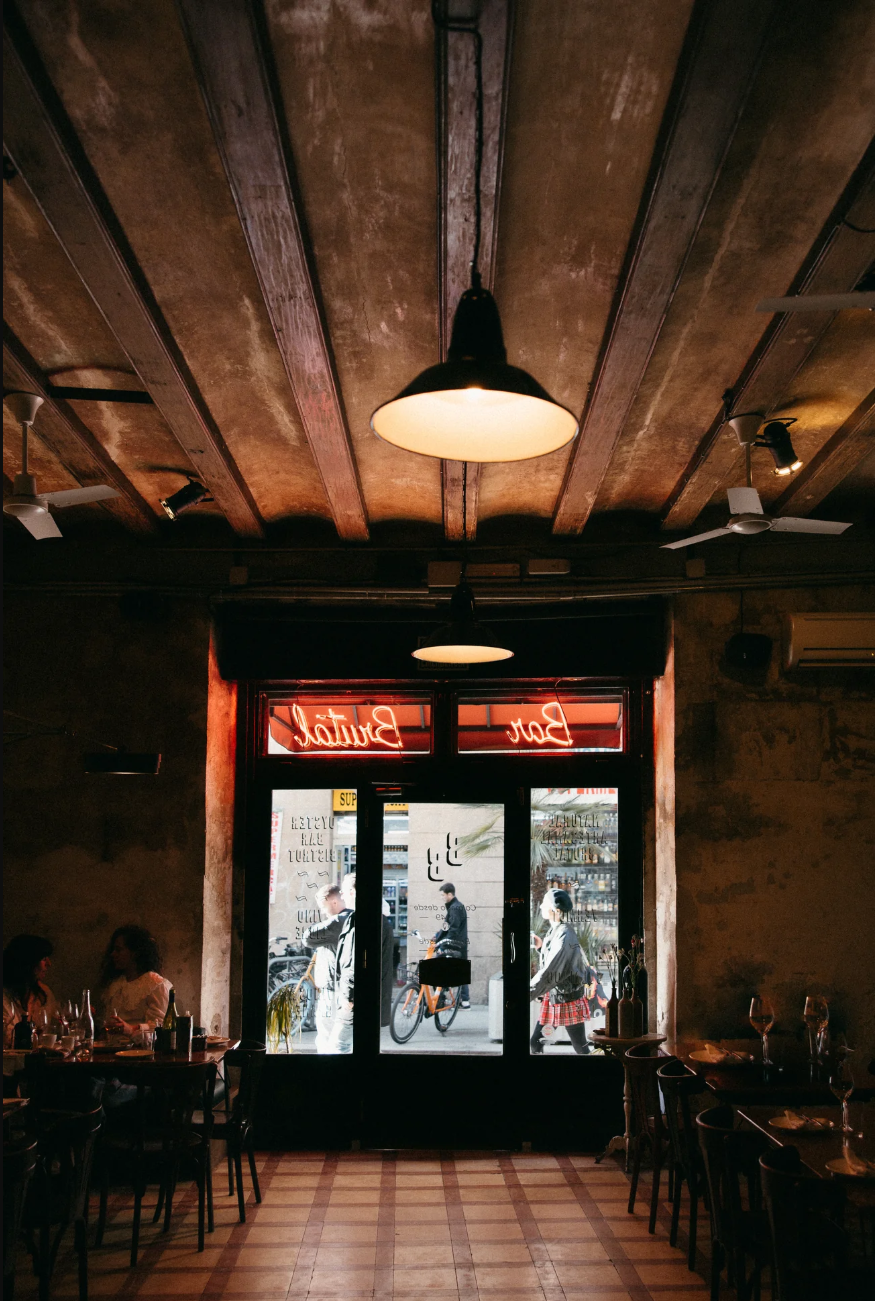 Interior of Bar Brutal featuring wooden ceiling beams, softly lit dining tables, and a glass entrance overlooking the street.