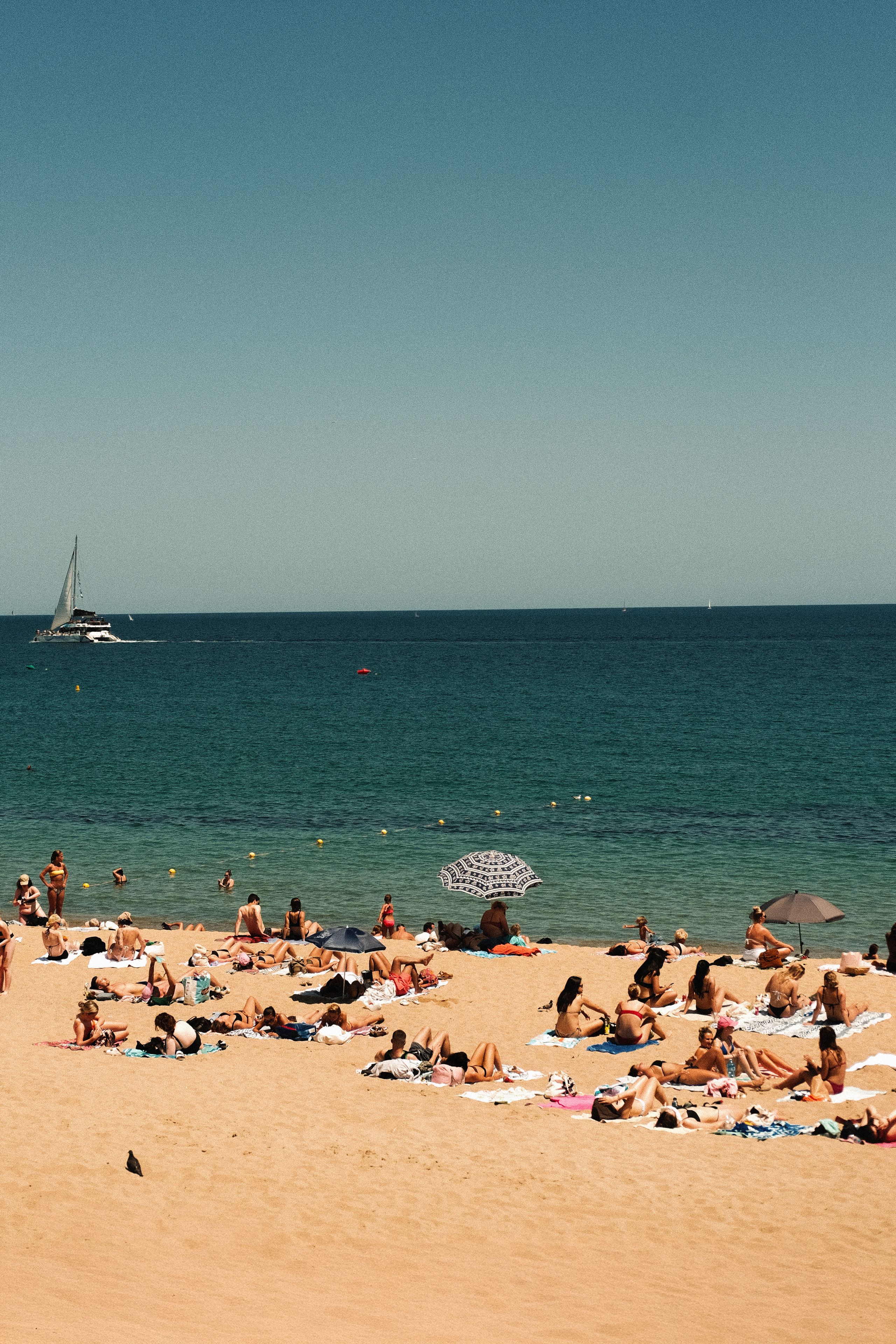 People sunbathing on Barceloneta Beach with the Mediterranean Sea and a sailboat in the background.